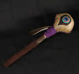 A large rattle with a peacock feather design, featuring a rawhide head and elk suede handle, embellished with copper wire.
