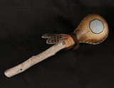 A large handheld rattle with a rawhide head, decorated with a painted geometric peyote design and feathers, with a suede leather wrapping and a wooden handle.