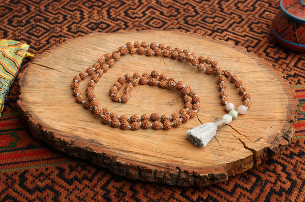 A mala necklace made of Rudraksha seeds, agate, and turquoise laid out on a wooden surface with a brahmagranti knot at the center and a white and blue tassel.